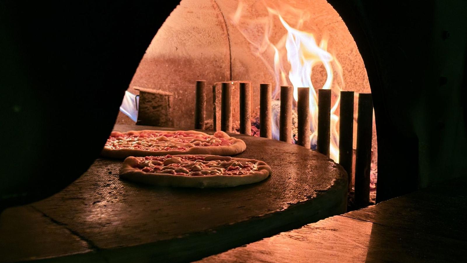 Two pizzas in the wood-fired oven with flames blazing behind them