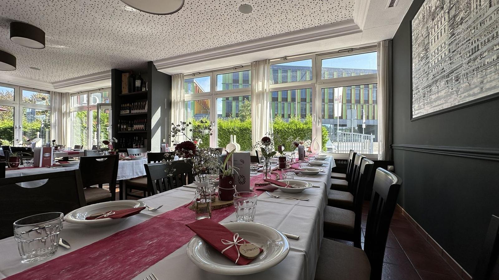 Heller Gastraum mit langer Hochzeitstafel, rotem Tischläufer und Blumen, durch die Fenster fällt Sonne