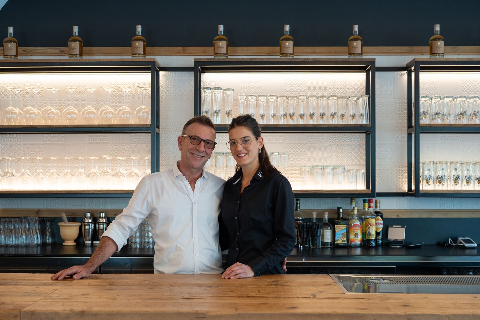 Serafino and Alesandra smiling behind the wooden counter, illuminated shelves of glasses behind them